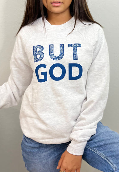 A girl is standing looking forward wearing a ash gray sweatshirt with big bold blue text that reads " But God". The word BUT on top of the word GOD. #color_ash-gray