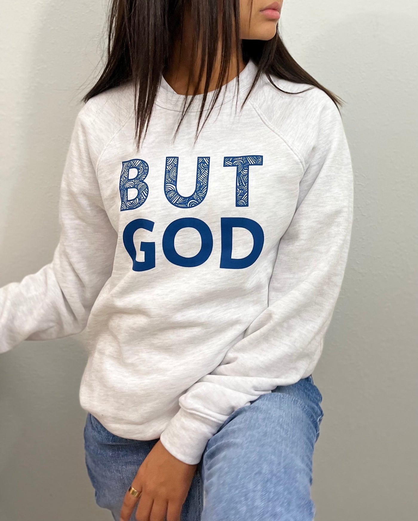 A girl is standing looking towards left wearing a ash gray sweatshirt with big bold blue text that reads " But God". The word BUT on top of  the word GOD. #color_ash-gray