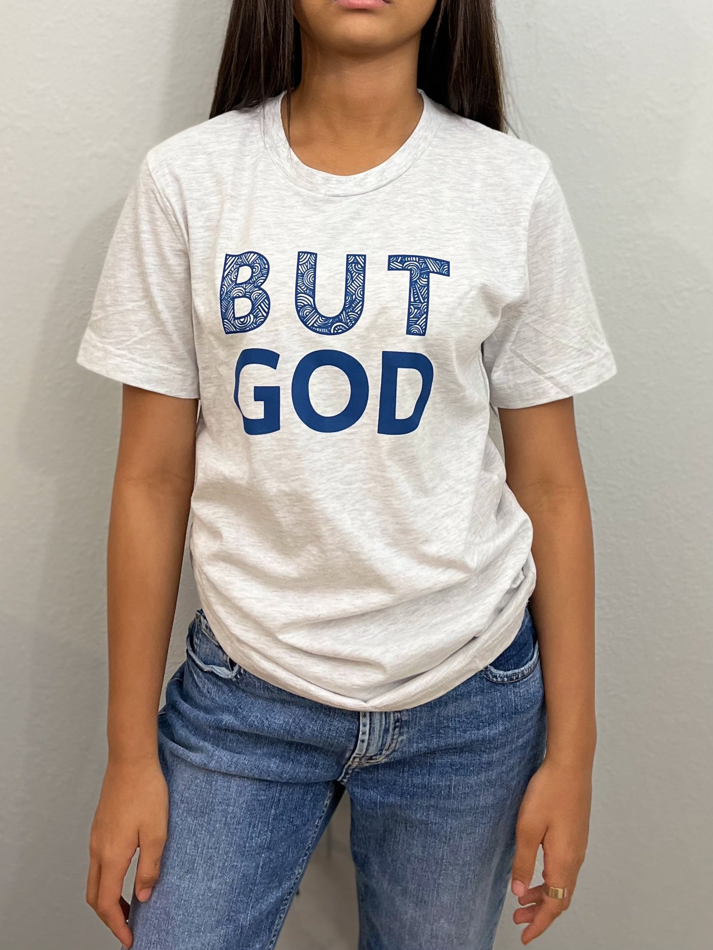 A girl is standing looking forward, wearing a ash gray t-shirt with big bold blue text that reads " But God". The word BUT on top of the word GOD. #color_ash-gray