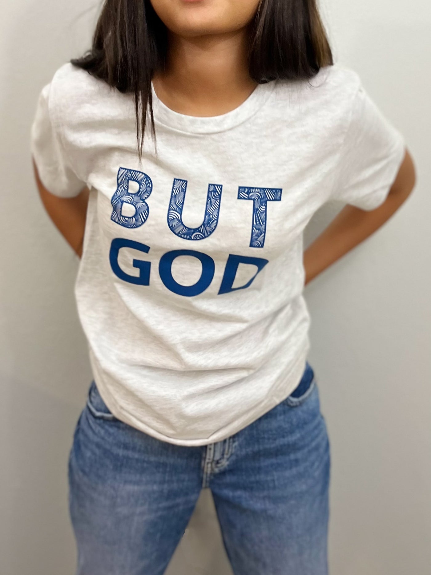 A girl in blue jeans is standing leaning forward, wearing a ash gray t-shirt with big bold blue text that reads " But God". The word BUT on top of the word GOD. #color_ash-gray
