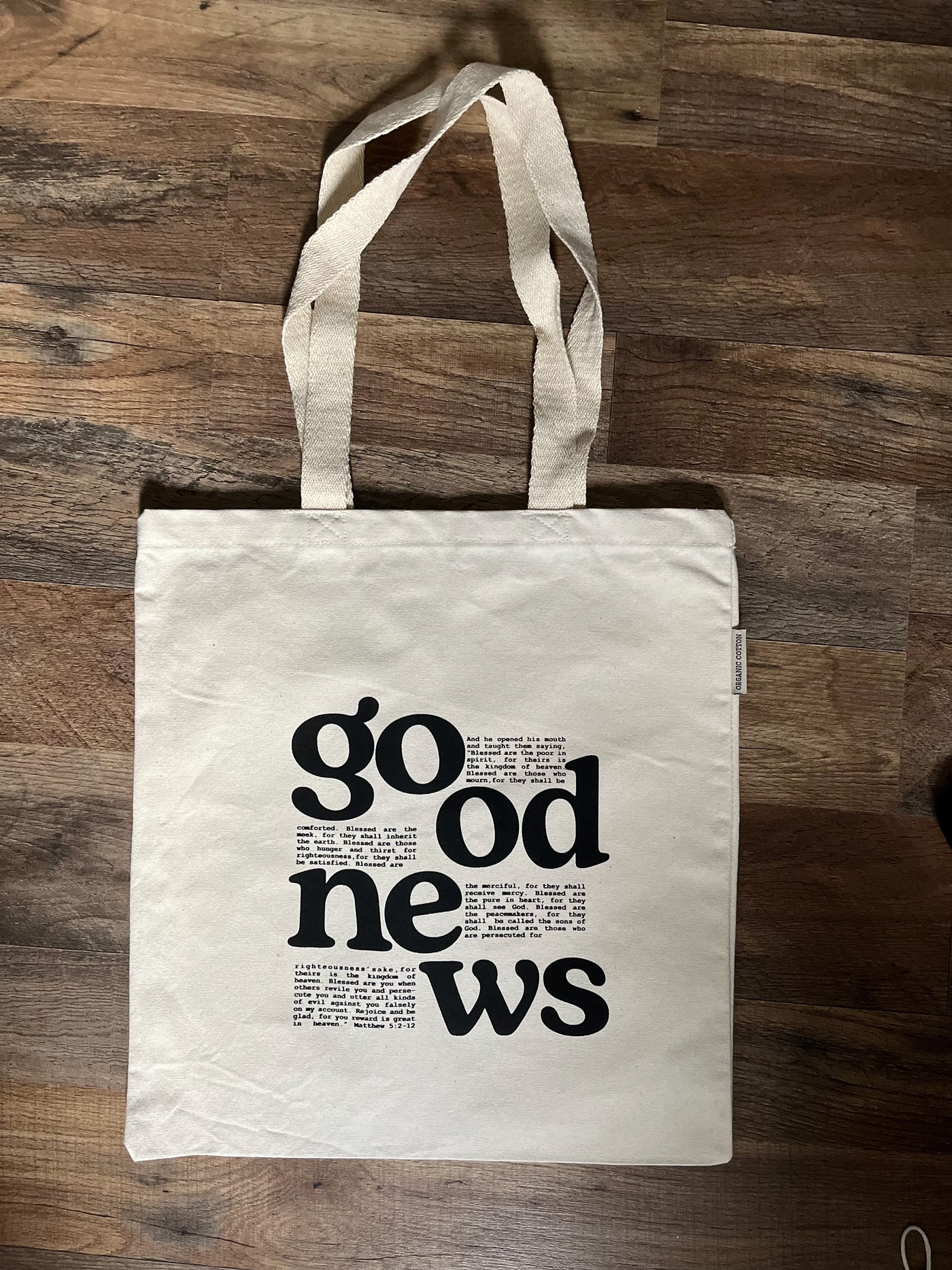 A tote bag is laying in the center of a wooden floor. The words good news are in bold font. Within the typography is a bible verse.