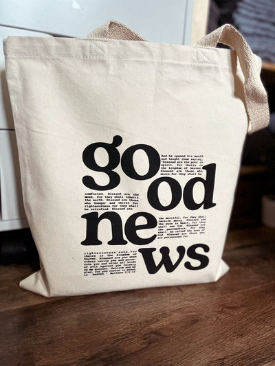 A natural canvas tote bag sits on a wooden floor, propped against a white dresser. The words good news are in bold font. Within the typography is a bible verse.