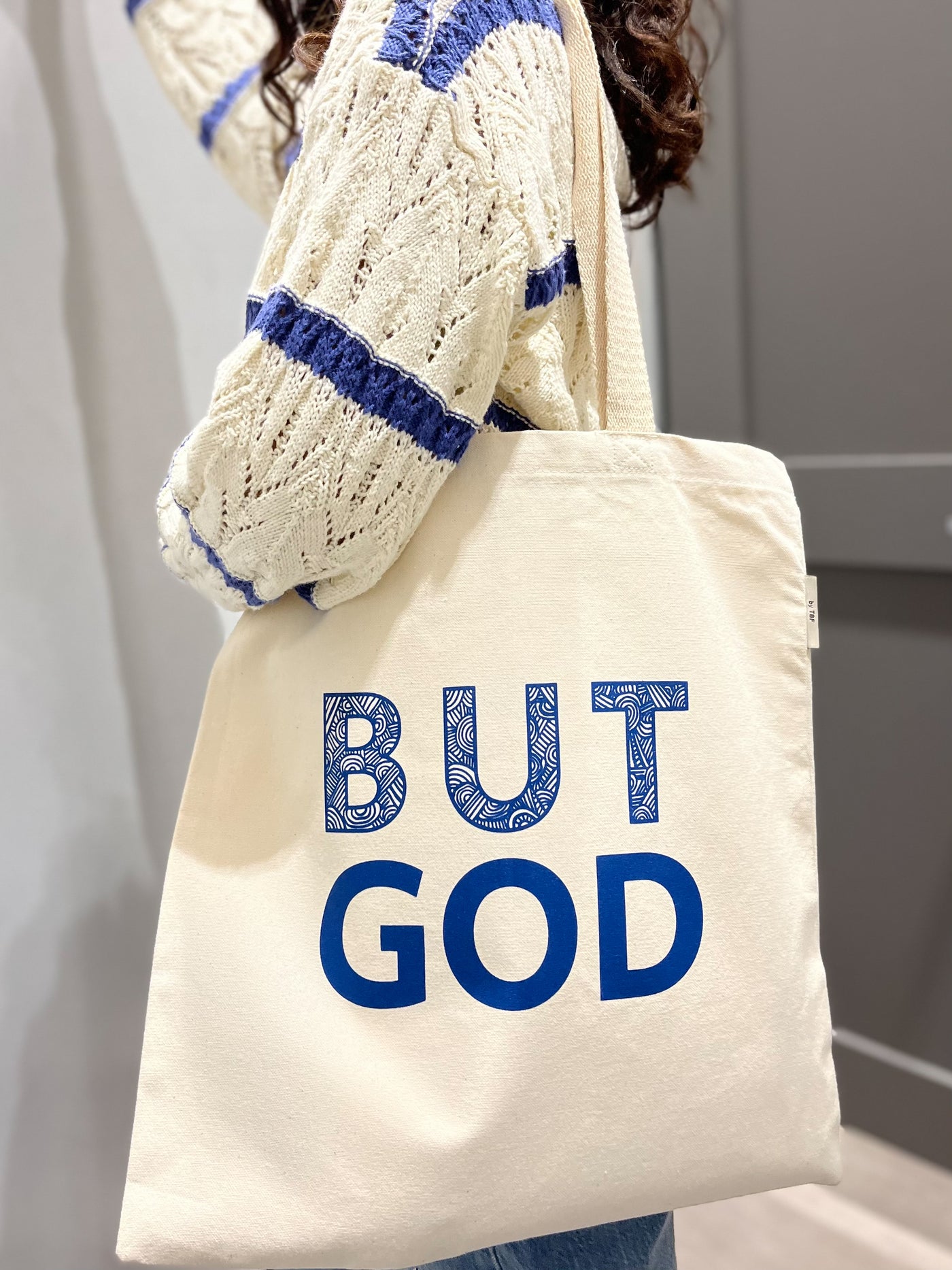 a girl is standing in bright room in front of a gray door. on her left side, she  holds a natural canvas tote bag with big blue text that reads  'BUT GOD". She has a blue and cream knit sweater on. the ends of her curly hair fall down her back.