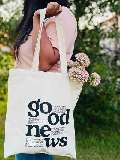 A woman standing in a grassy outside holding a  natural canvas tote bag in her left hand. The tote bag reads good news and displays a bible verse in between the typography. Pink flowers are hanging out of the side of the bag towards the back.